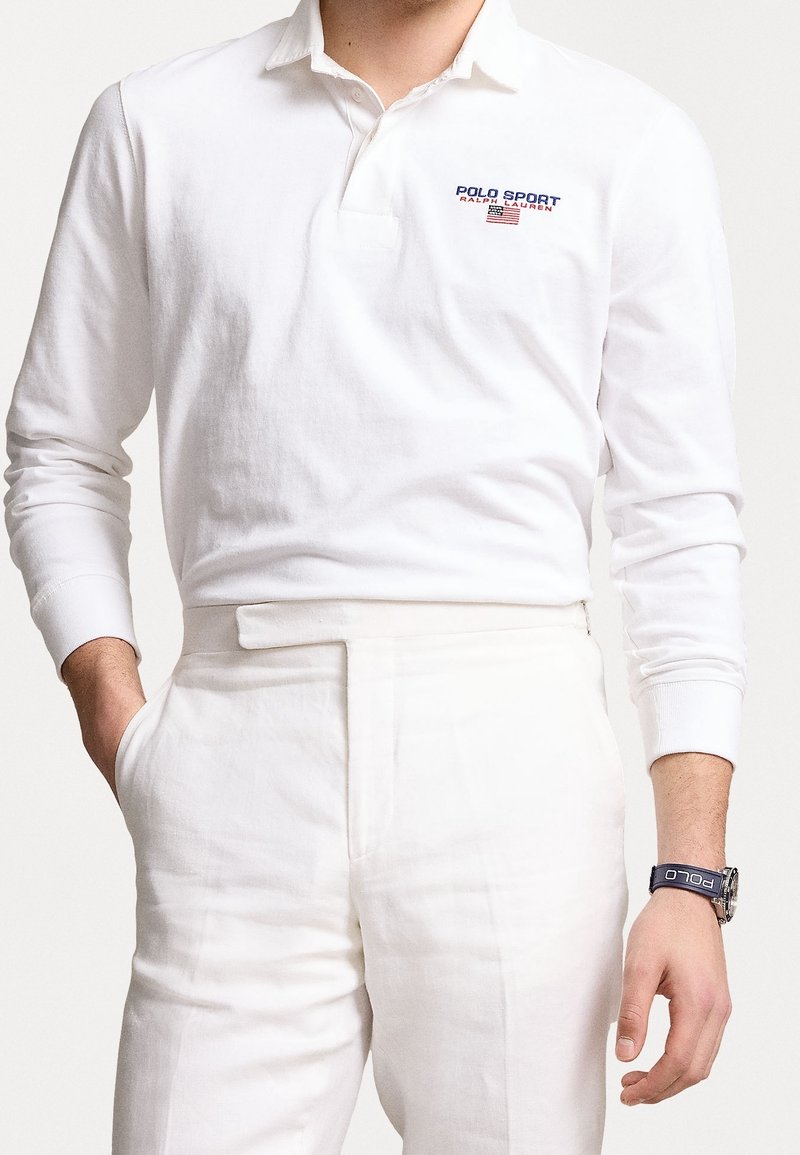 Vit långärmad pikétröja med knäppt krage och blå/röd brodering av "POLO SPORT". Kombinerad med ljusa byxor och en klocka.