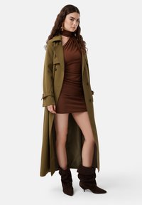 Trench coat verde oliva sopra un abito aderente marrone, caratterizzato da un collo alto e una texture morbida. Stivali alla caviglia marroni morbidi completano il look.