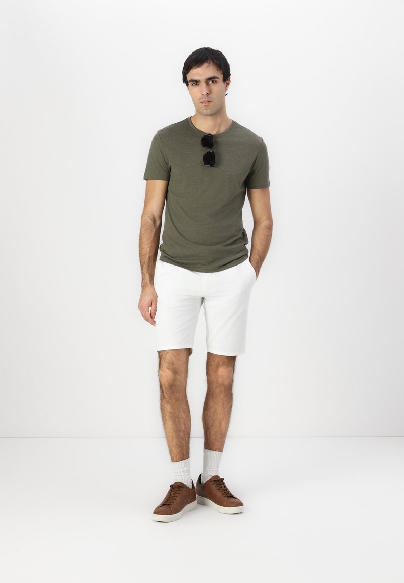 Olivgrünes T-Shirt, weiße Shorts, braune Sneaker und weiße Socken. Sonnenbrille auf dem T-Shirt platziert. Modell steht vor einem weißen Hintergrund.
