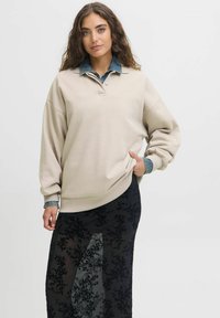 Beige oversized sweatshirt with a collared neckline, layered over a sheer black skirt featuring floral patterns.