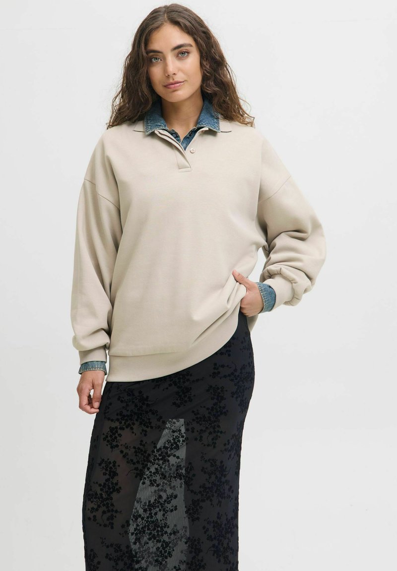 Beige oversized sweatshirt with a collared neckline, layered over a sheer black skirt featuring floral patterns.