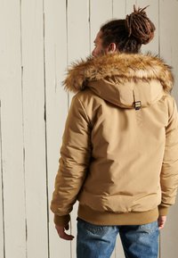 Chaqueta de plumas marrón claro con capucha forrada de piel sintética, puños y dobladillo acanalados. Textura suave con un diseño ceñido y detalle de cierre en la parte trasera.