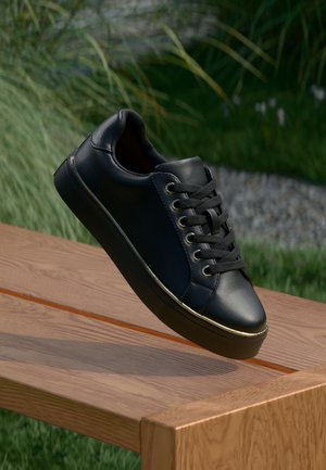 Schwarzer Ledersneaker mit schwarzen Schnürsenkeln und goldenen Ösen schwebt über einer Holzbank im Freien, mit Gras und Pflanzen im Hintergrund.