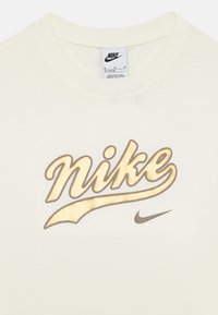 Kremkrāsas kokvilnas T-krekls ar zelta un brūna stilizētu "Nike" logotipu un mazu brūnu Swoosh. Apaļa kakla izgriezne.