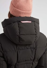 Chaqueta de plumas negra con cuello alto y textura acolchada. Presenta una etiqueta rosa en la parte posterior, complementada por un gorro de punto rosa claro.