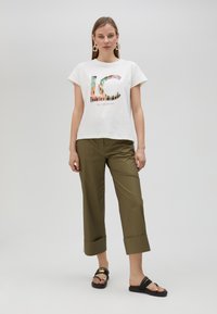 Camiseta de algodón blanca con un gráfico colorido de "LC" en el frente, combinada con pantalones cortos de color verde oliva y sandalias negras.