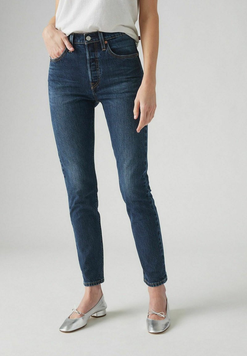 Vrouw in donkerblauwe hoge taille skinny jeans, witte top en zilveren lage ballerina's, staand met één hand in de zak.
