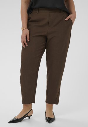 Žena v krátkych hnedých nohaviciach a čiernych nízkych lodičkách na pätke s otvorenou pätou, s jednou rukou v vrecku a zlatým náramkom na zápästí.