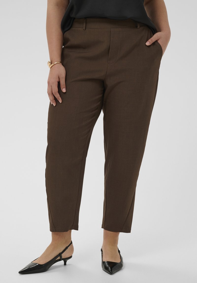 Femme portant un pantalon marron court et des chaussures slingback noires à petit talon, avec une main dans la poche et un bracelet doré au poignet.