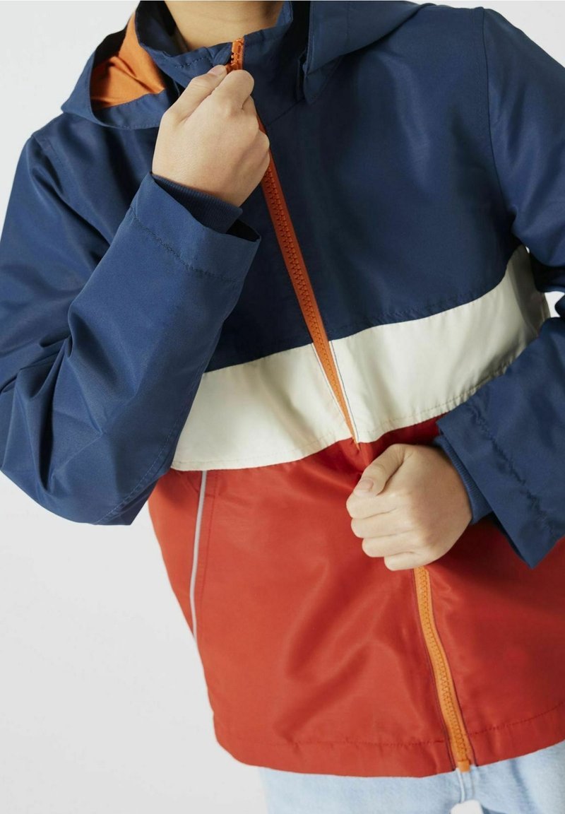 Veste colorée avec une partie supérieure bleu marine, une bande centrale blanche et un bas rouge. Dotée d'une fermeture à glissière et d'une capuche réglable.