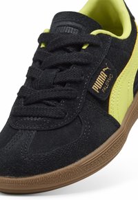 Zwarte suède sneakers met een gele accent en een bruine rubberen zool. Voorzien van platte veters, een gevoerde kraag en een merklabel aan de zijkant.