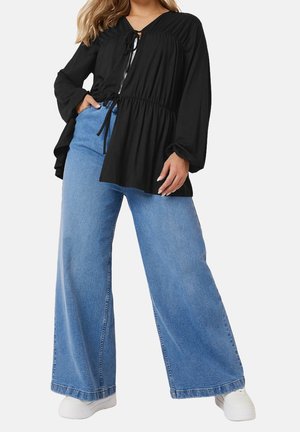 Kvinde iført løs sort bluse med lange ærmer og vidde blå jeans med hvide sneakers, stående med den ene hånd i lommen.