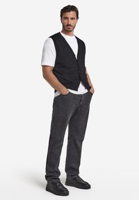 Schwarze Strickweste über einem weißen T-Shirt, dunkelgraue Jeans und schwarze Schuhe. Die Weste hat einen Knopfverschluss und eine lockere Passform.