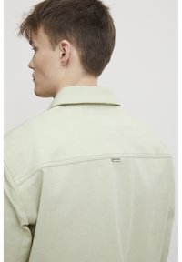 Lys grøn skjorte med krave laget av mykt stoff, med et merke på baksiden og et enkelt design med minimale sømdetaljer.