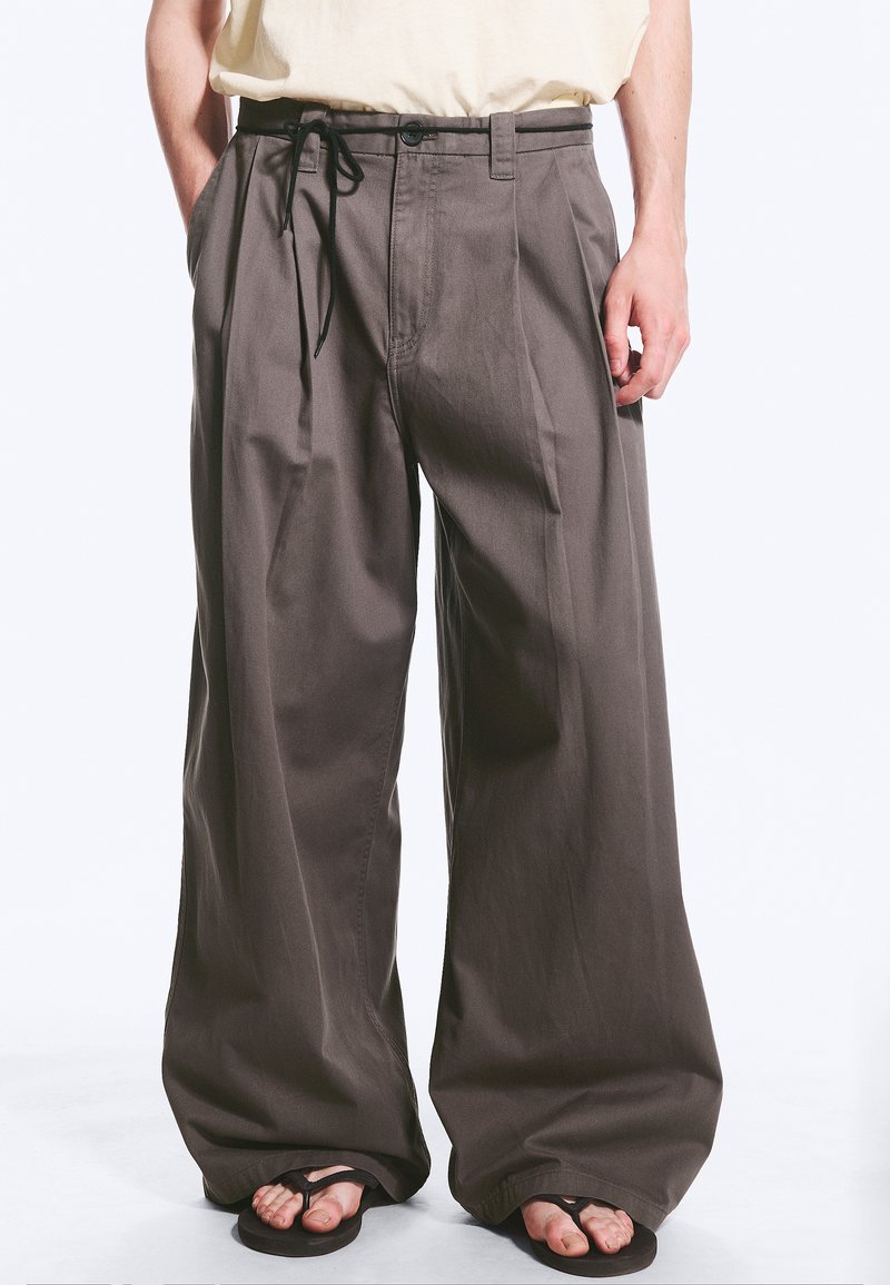 Homme portant un pantalon marron à jambes larges, une chemise claire rentrée, et des tongs noires avec les orteils visibles.