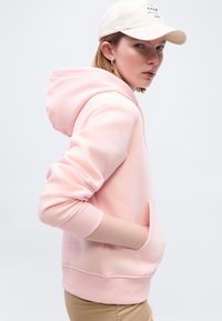 Νεαρό άτομο με ανοιχτό ροζ φούτερ με κουκούλα, με τα χέρια στις τσέπες, φορώντας μπεζ καπέλο και χρυσό σκουλαρίκι κρίκο, κοιτάζοντας πλάγια σε ένα απλό φόντο.