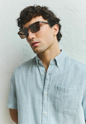 Vīrietis ar cirtainiem tumšiem matiem, valkājot pārāk lielas kvadrātveida tumšas saulesbrilles un gaiši zilu pogu kreklu, stāv pret teksturētu balto sienu.