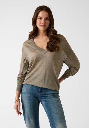 Kobieta o długich brązowych włosach, ubrana w luźny beżowy sweter z dekoltem w serek i niebieskie jeansy, stojąca z jedną ręką na biodrze na białym tle.