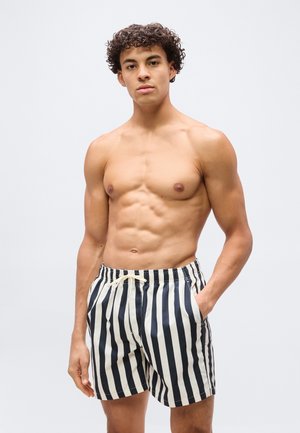 Joven con cabello rizado que lleva pantalones cortos de baño con rayas verticales en blanco y negro, de pie con una mano en el bolsillo contra un fondo liso.