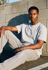 Ung mann i lysegrå T-skjorte og bukser som sitter på bakken, lent mot en betongblokkvegg i sollys, ser rolig framover.