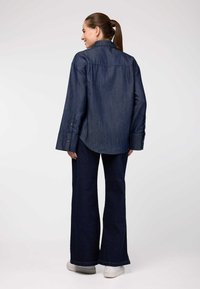 Camicia in denim blu scuro a maniche lunghe, con polsini abbottonati e orlo curvo, indossata con pantaloni svasati scuri abbinati e sneakers bianche.