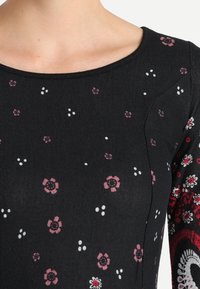 Haut en tissu noir avec un motif floral en rose et blanc, col rond, manches texturées et accents de motif sur les côtés.
