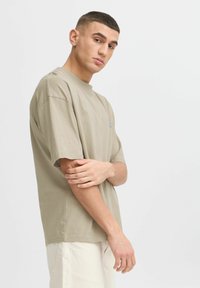 T-shirt à manches courtes, couleur beige clair, en coton doux avec un col rond. Il présente un petit motif bleu sur la poitrine gauche et a une coupe décontractée.