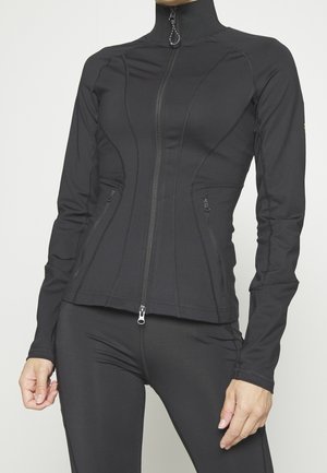 Kobieta ubrana w dopasowaną czarną kurtkę zapinaną na zamek z wysokim kołnierzem i dopasowane czarne legginsy na jednolitym tle.