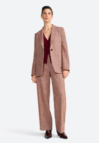 Kudottu tweed-puku vaaleanruskeana ja ruosteenvärisenä, jossa on jämäkkä bleiseri yhdellä napilla ja leveälahkeiset housut. Aluskerros on burgundin värinen.