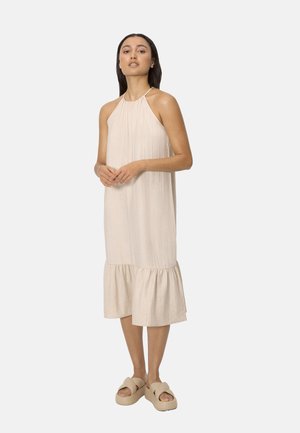 Mujer con vestido midi sin mangas beige con dobladillo fruncido y escote halter, combinado con sandalias plataforma beige, de pie.