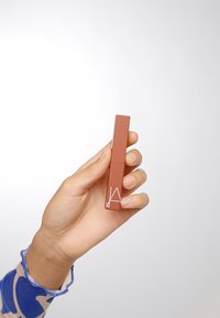 Un rouge à lèvres liquide dans un emballage rectangulaire en rose poussiéreux, avec un design épuré et un logo blanc. Tenue dans une main avec un teint clair.