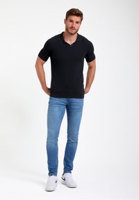 Melnā īsā piedurkņu polo krekls ar teksturētu audumu, ar apkakli un taisnu apakšmalu. Nēsāts ar gaiši ziliem džinsiem un baltiem sporta apaviem.