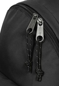Schwarzer Stoff-Rucksack mit doppelten silbernen Reißverschlüssen, die texturierte Ziehungen und geflochtene schwarze Schnüre aufweisen. Auffällige Nähedetails auf dem Stoff.