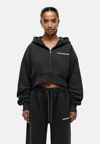 Felpa cropped nera con zip, maniche oversize e logo bianco "GOODFORNOTHING"; abbinata a pantaloni della tuta neri con cintura elastica.