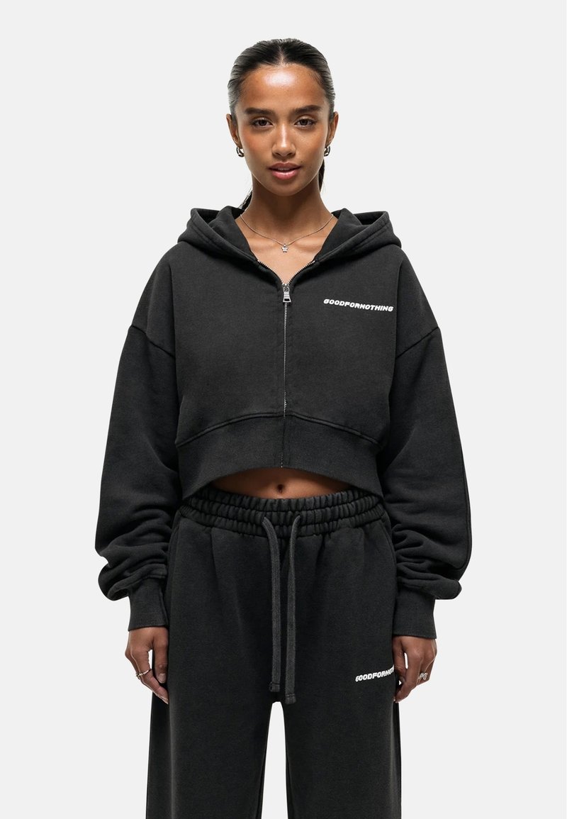 Felpa cropped nera con zip, maniche oversize e logo bianco "GOODFORNOTHING"; abbinata a pantaloni della tuta neri con cintura elastica.