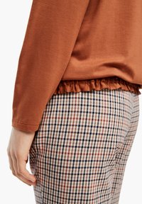 Rdzawy top z falbaniastym dołem w połączeniu z woskowanymi spodniami w neutralnych odcieniach z czarnymi, brązowymi i czerwonymi liniami. Gładka tekstura materiału.