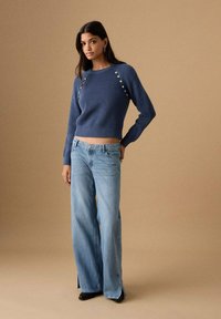 Navy plavi pleteni džemper s okruglim izrezom i ukrasnim gumbima na ramenima, uparen s lagano plavim trapericama širokih nogavica i crnim cipelama. Jednostavne teksture.