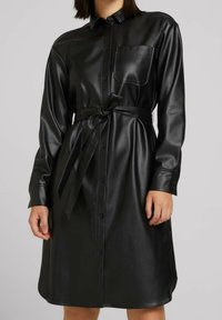 Schwarzes Kleid aus Kunstleder mit durchgehender Knopfleiste, langen Ärmeln, Brusttasche und gebundenem Gürtel in der Taille. Glatte Textur.