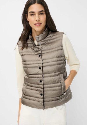 Frau trägt taupe farbene, gesteppte ärmellose Steppweste mit Druckknöpfen über einem weißen Langarmshirt, Hand in der Westentasche, neutraler Hintergrund.