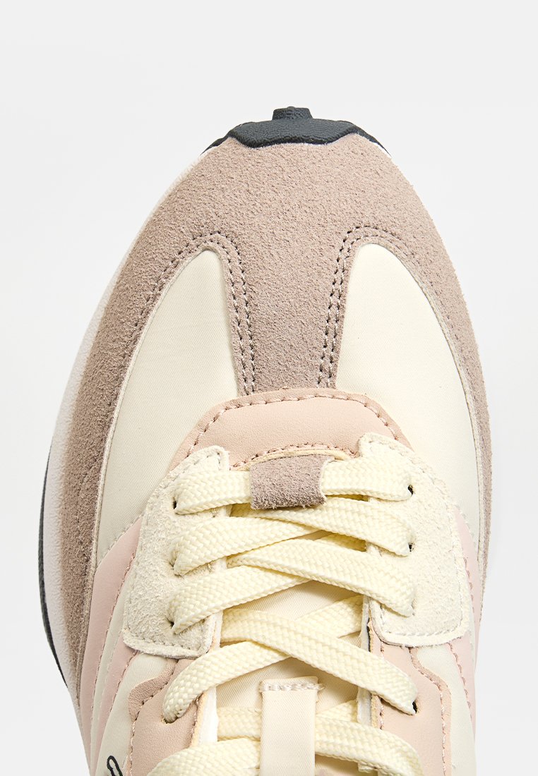 Zapatilla con una parte superior de color crema, suaves acentos de ante beige y suela de goma negra texturizada. Presenta cordones planos color marfil y costuras sutiles.