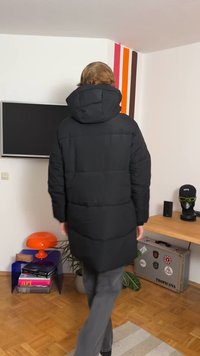 Juoda pūkinė striukė su gaubtu, pasižyminti siūta faktūra, ilgo kirpimo ir elastinėmis rankogaliais. Dėvima su pilkomis kelnėmis moderniame interjere.