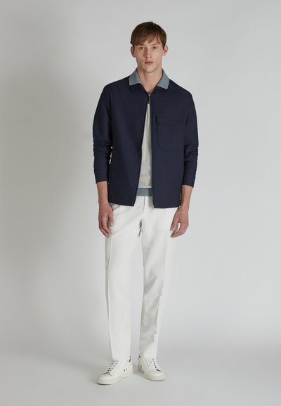 Veste zippée marine avec un col bleu clair, en tissu texturé, détails de poches, associée à un pantalon blanc et des baskets blanches.