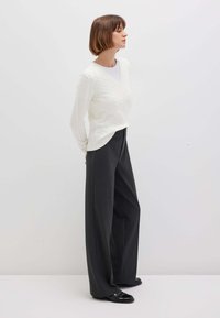 Donna in piedi di lato, che indossa un maglione bianco a treccia, pantaloni larghi scuri e mocassini neri su uno sfondo neutro.