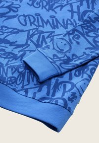 Manica di felpa blu con testo in stile graffiti blu scuro e motivi di faccine sorridenti, polsini e orlo a coste su uno sfondo chiaro.