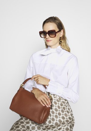 Mala de mão em couro castanho com acabamento texturizado, ferragens douradas e uma alça curta. Usada com uma blusa branca com detalhe em laço.