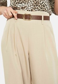 Bēša augsta vidukļa bikses ar krokiem un brūnu ādas jostu ar zelta sprādzi, kombinētas ar leoparda raksta topu.