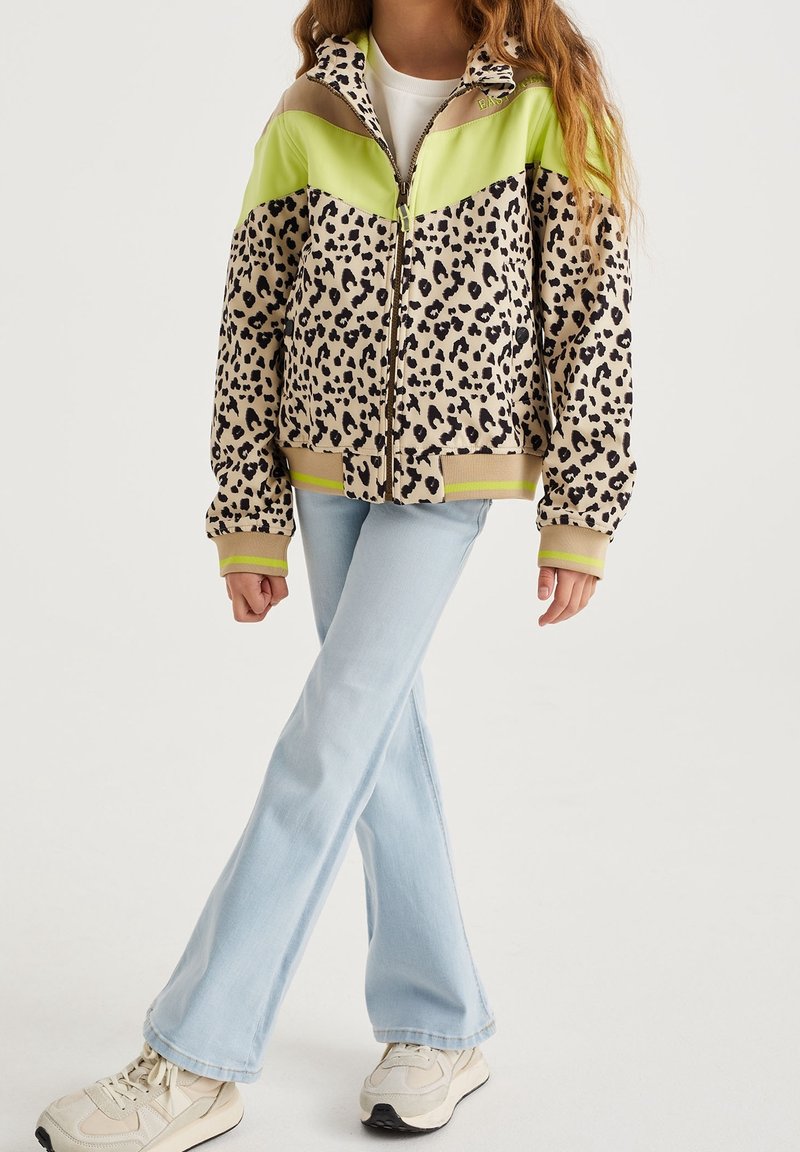 Bambino che indossa una giacca zip-up con stampa leopardata e gialla neon, jeans a zampa d'elefante blu chiaro e sneaker beige, in piedi con le gambe incrociate.