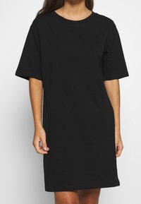 Nainen, joka on pukeutunut yksinkertaiseen mustaan lyhythihaiseen T-paitamekkoon ja seisoo valkoista taustaa vasten.