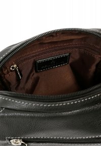 Interno di una borsa in pelle nera con una texture liscia, foderato in tessuto marrone, con una tasca con zip e un piccolo tag con logo.