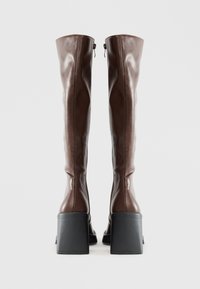 Bottes en cuir marron montantes avec une finition lisse, des talons épais noirs et une fermeture éclair à l'arrière.